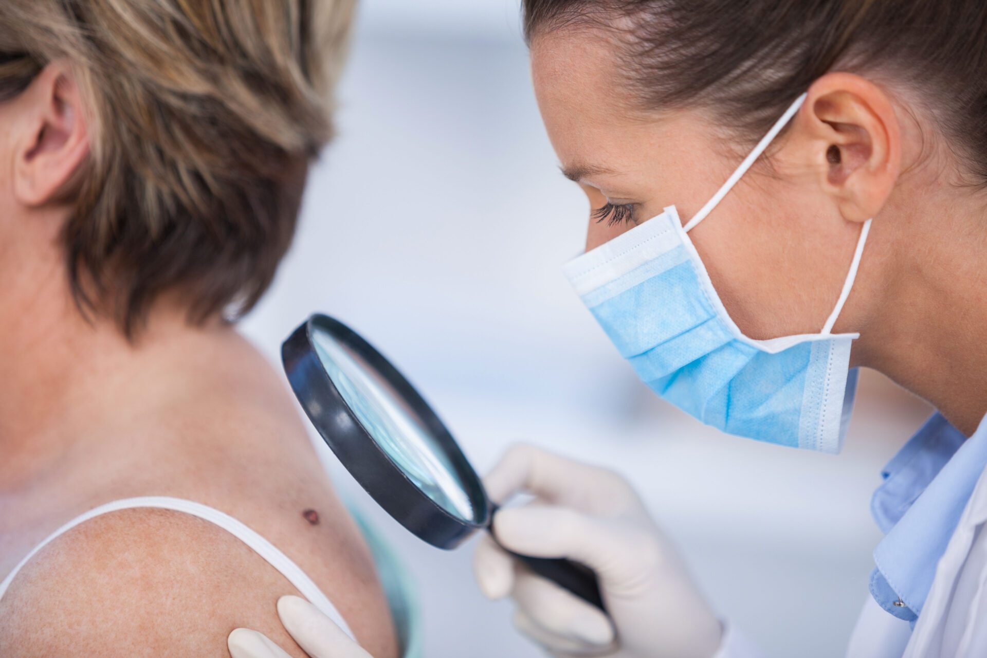 Dermatologist examining mole of female patient with magnifying glass in clinic